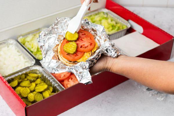 A close-up photo of a person putting pickles on their cheeseburger in front of a Five Guys catering box of toppings.