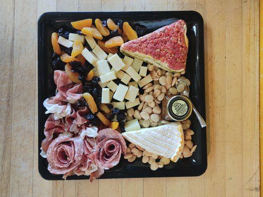 A photo of a square charcuterie board featuring Melody's Sun-Dried Tomato & Pesto Torta, Vacherousse, Genoa Salami, Prosciutto, and Cheddar.