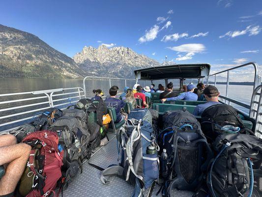 A group of people on a boat, facing away from the camera. Mountains are in the background and 13 backpacking backpacks are in the foreground