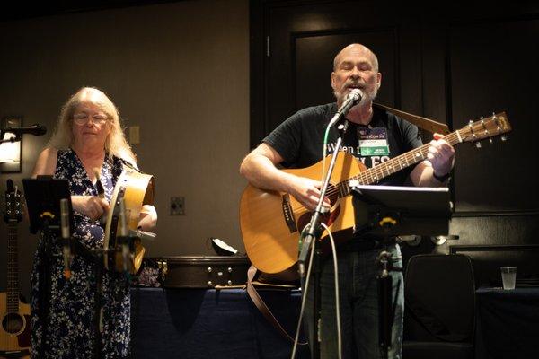 Two musicians, one with long white hair and a drum, one with a beard and a guitar.