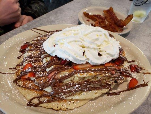 Tall stack of Chocolate chip pancakes; "Cloud 9" style (whipped cream, Nutella, and strawberries added), with a side of bacon.