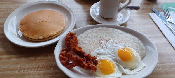 Short stack of pancakes,  sunnyside up eggs, bacon and cheese grits as presented