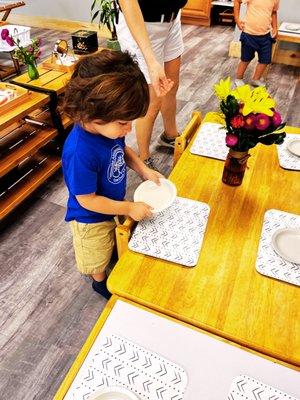 Child setting the table for classmates during lunch. Practical life exercises instill care for self, others and the environment.