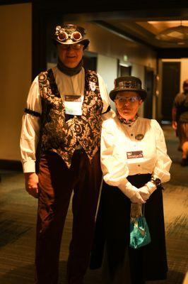 A couple, one in top hat and embroidered vest, one in top hat, poofy white top and dark skirt, with a green handbag.