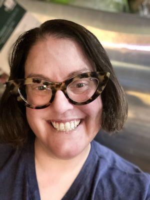 A white-passing woman with dark brown hair wearing cat-eye tortoiseshell glasses and a blue shirt.