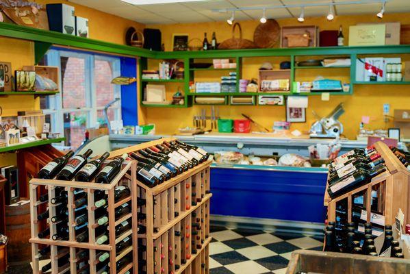 From the entrance, full wine racks, a cheese and deli case, and gift items on shelves.