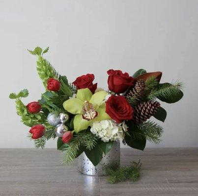 A shimmering arrangement of hydrangea, red roses, and a stunning cymbidium orchid with holiday accents such as ornaments and pinecones.