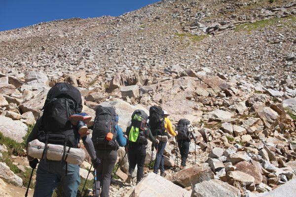 5 hikers hiking on a trail, walking away from the photographer with a scree field (rock debris field) in the background