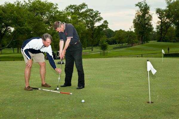 A volunteer helping a blind man line up a golf putt.