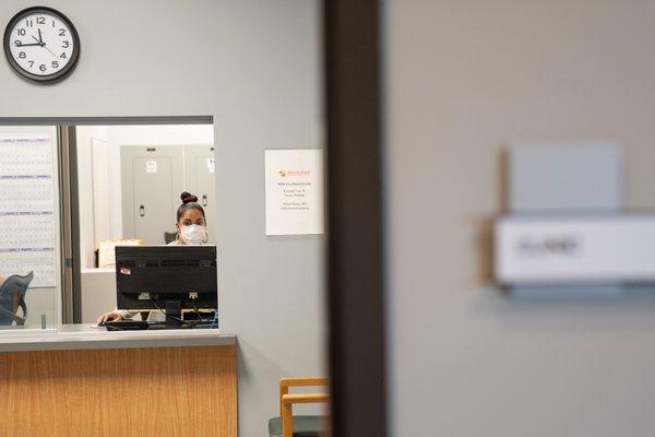 An open doorway shows a waiting room and a reception window with a woman sitting at a computer