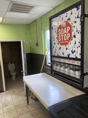 Laundry detergent vending machine hovers over folding table, located just outside of unisex bathroom.