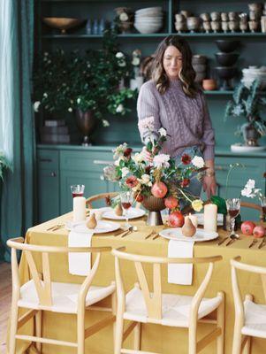 floral designer creating tablescape in flower studio. green painted walls, golden table linen, florals and fruit