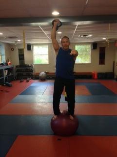 Gary standing on a stability ball with a kettlebell overhead.